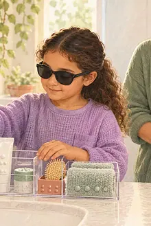 A blind child organizes toiletries at a sink while a caregiver waits nearby