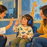 A pediatric audiologist giving a young child a playful hearing test while a parent watches