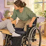 A teenager and caregiver coordinate a transfer between a bed and wheelchair using a transfer board